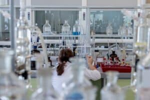 En person i vit labbrock sitter vid en laboratoriebänk med glasflaskor och provrör.